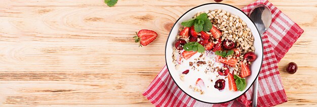 Desayuno saludable - granola, fresas, cerezas, nueces y yogurt en un recipiente sobre una mesa de madera. Concepto de comida vegetariana. Vista superior