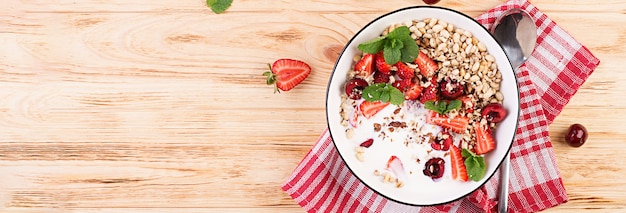 Desayuno saludable - granola, fresas, cerezas, nueces y yogurt en un recipiente sobre una mesa de madera. Concepto de comida vegetariana. Vista superior