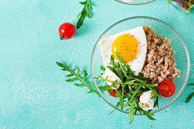 Desayuno saludable con huevo, queso feta, rúcula, tomate y gachas de trigo sarraceno sobre fondo claro. Nutrición apropiada. Menú dietético Endecha plana. Vista superior