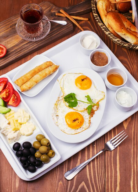 Desayuno turco con huevos fritos, tomate, pepino, quesos, aceitunas negras, miel, mermelada, queso crema, pan galeta y vaso de té.