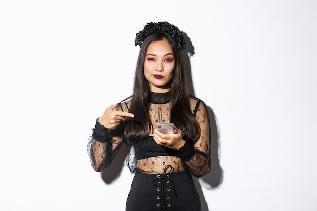 Descarada hermosa mujer asiática en vestido gótico y corona negra apuntando con el dedo en el teléfono móvil, mostrando algo sobre halloween.
