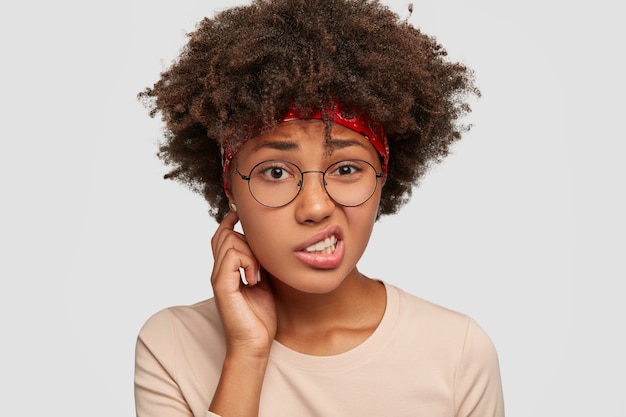 Foto gratuita desconcertada e incierta niña negra frunce el ceño con disgusto, siente apatía, mira con incertidumbre, no encuentra solución y salida del problema, tiene corte de pelo afro, modelos contra la pared blanca. expresión facial