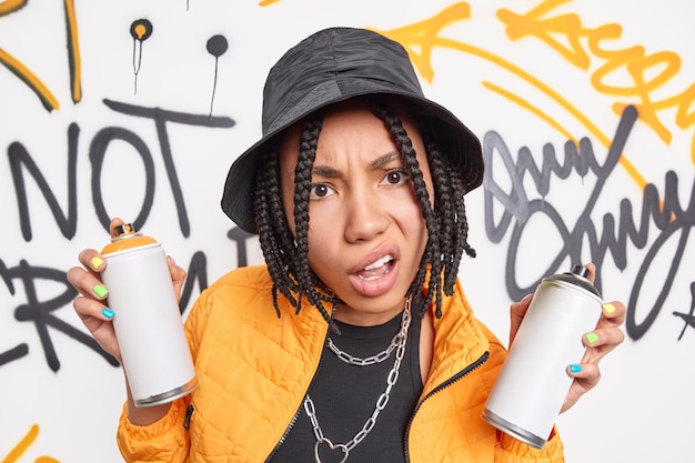 Descontento adolescente con trenzas sostiene latas de aerosol para hacer graffiti vestida con chaqueta amarilla, sombrero negro, cadenas de metal alrededor del cuello, disfruta de la cara de sonrisas de estilo urbano