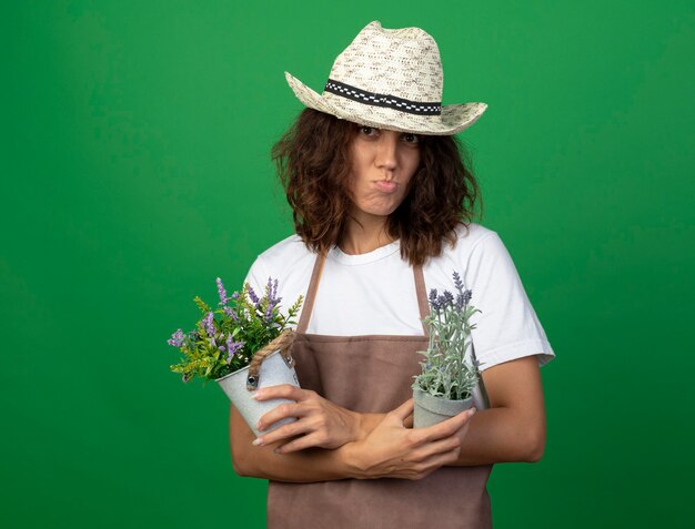 Descontento jardinero de sexo femenino joven en uniforme vistiendo sombrero de jardinería sosteniendo y cruzando flores en macetas