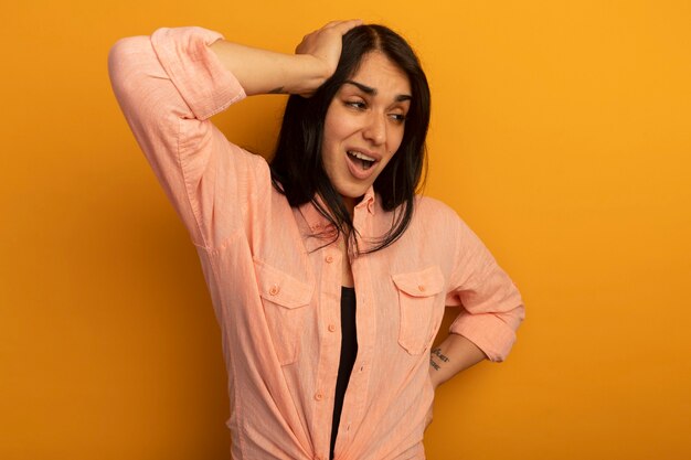 Descontento mirando al lado hermosa joven vestida con camiseta rosa poniendo las manos en la cadera y la cabeza aislada en amarillo