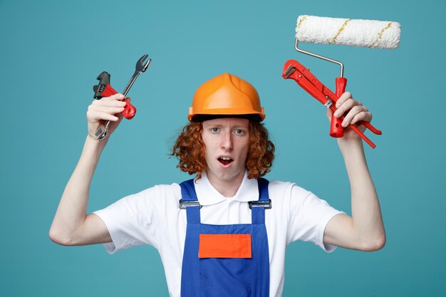 Descontento mirando a la cámara joven constructor hombre en uniforme sosteniendo herramientas de construcción aislado sobre fondo azul.