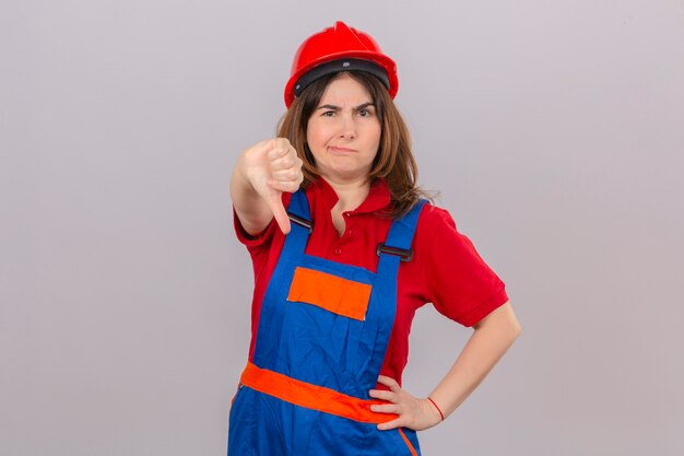 Descontentó a la mujer constructora con uniforme de construcción y casco de seguridad mostrando el pulgar hacia abajo con el ceño fruncido de pie sobre la pared blanca