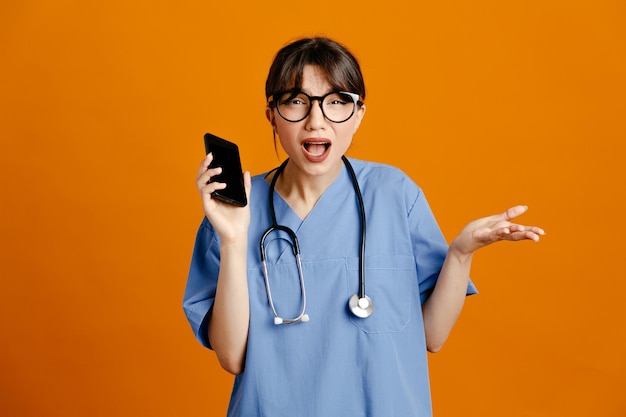 Descontento sosteniendo teléfono joven doctora vistiendo uniforme fith estetoscopio aislado sobre fondo naranja