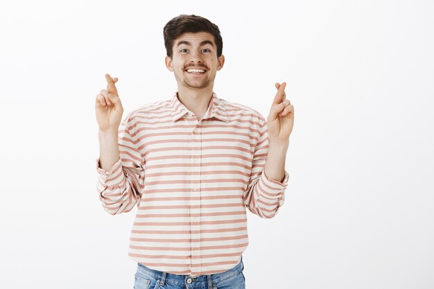 Deséame suerte. Retrato de sonriente amable modelo masculino maduro con bigote, cruzando los dedos y sonriendo ampliamente, esperando o rezando por un negocio exitoso, de pie