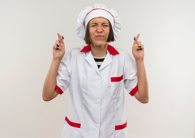 Deseando joven cocinera en uniforme de chef con los dedos cruzados y los ojos cerrados aislado sobre fondo blanco con espacio de copia
