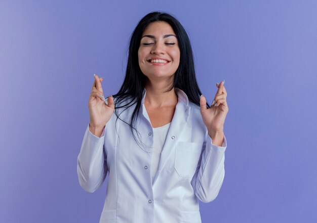 Deseando joven doctora vistiendo bata médica haciendo gesto de dedos cruzados con los ojos cerrados