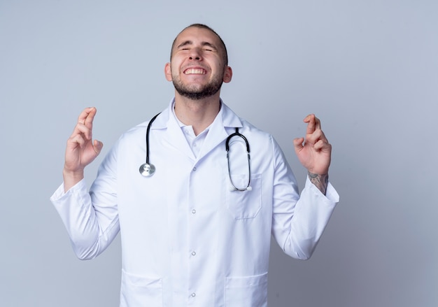 Deseando joven médico con bata médica y estetoscopio alrededor de su cuello haciendo gesto de dedos cruzados con los ojos cerrados aislado sobre fondo blanco con espacio de copia