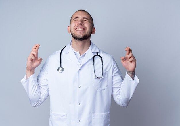 Deseando joven médico con bata médica y estetoscopio alrededor de su cuello haciendo gesto de dedos cruzados con los ojos cerrados aislado sobre fondo blanco con espacio de copia