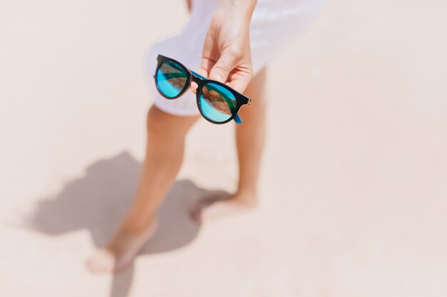 Desenfoque de retrato de dama bien formada de pie sobre la arena con vestido blanco. Tiro al aire libre de mujer bronceada caucásica escalofriante en la playa y con gafas de sol.
