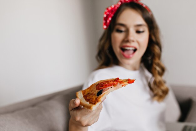 Desenfoque de retrato de mujer joven alegre mirando rebanada de pizza. Foto interior de divertida chica relajante disfrutando de comida rápida en casa.