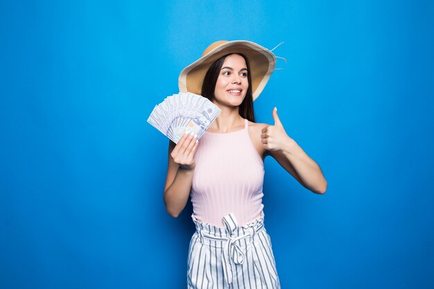 Desgaste atractivo de la mujer en el sombrero de paja que muestra los billetes de 100 usd, pulgar hacia arriba, aislado sobre la pared azul.