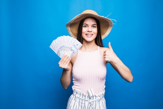 Desgaste atractivo de la mujer en el sombrero de paja que muestra los billetes de 100 usd, pulgar hacia arriba, aislado sobre la pared azul.