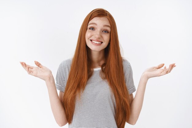 Desorientada encantadora tierna pelirroja de ojos azules niña sonriente inconsciente encogiéndose de hombros con las manos extendidas hacia los lados no tiene ni idea cuestionado disculparse no puede responder, de pie dudosa pared blanca