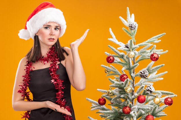 Desorientada joven bastante caucásica vestida con gorro de Papá Noel y guirnalda de oropel alrededor del cuello de pie cerca del árbol de Navidad decorado apuntando a él aislado en la pared naranja