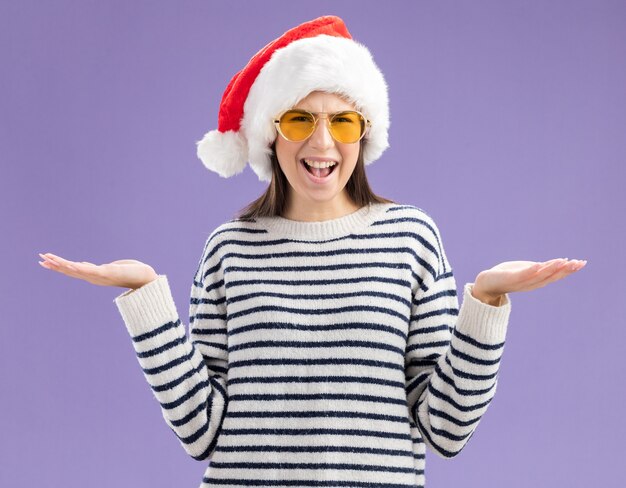 Desorientada joven caucásica en gafas de sol con gorro de Papá Noel sosteniendo las manos abiertas aisladas en la pared púrpura con espacio de copia