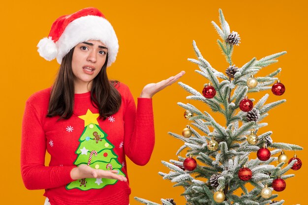Desorientada joven caucásica con gorro de Papá Noel apuntando al árbol de Navidad aislado sobre fondo naranja con espacio de copia