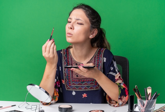 Desorientada joven morena sentada a la mesa con herramientas de maquillaje sosteniendo la paleta de sombra de ojos y mirando el pincel de maquillaje