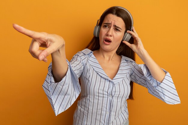 Desorientada mujer bastante caucásica en auriculares mira y apunta al lado en naranja