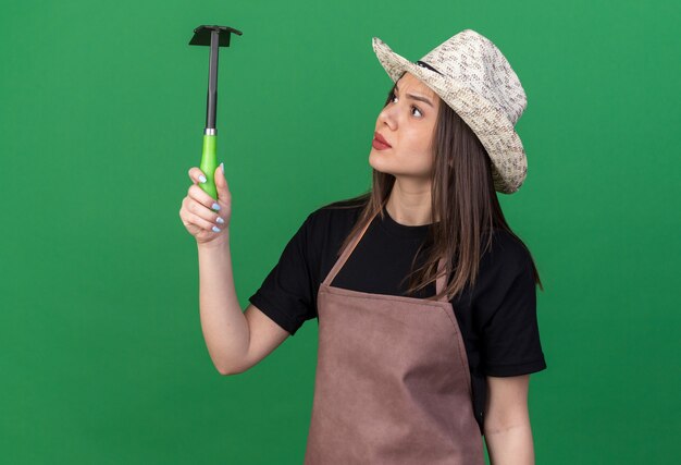 Desorientado bastante jardinero mujer caucásica vistiendo sombrero de jardinería sosteniendo y mirando azada rastrillo