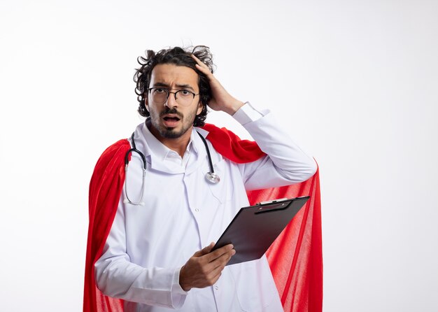 Desorientado, joven, caucásico, superhéroe, hombre, en, anteojos ópticos, llevando, doctor, uniforme, con, capa roja, y, con, estetoscopio, alrededor, cuello, pone, mano, cabeza, y, tenencia, portapapeles, con, espacio de copia