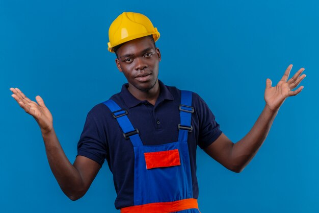 Desorientado joven constructor afroamericano hombre vestido con uniforme de construcción y casco de seguridad encogiéndose de hombros mirando incierto y confundido sin respuesta extendiendo las palmas de pie