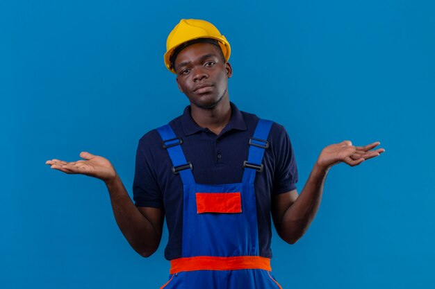 Desorientado joven constructor afroamericano hombre vestido con uniforme de construcción y casco de seguridad encogiéndose de hombros mirando incierto y confundido sin respuesta extendiendo las palmas de pie