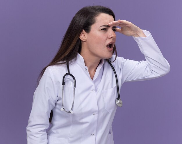 Desorientado joven doctora vistiendo bata médica con estetoscopio mantiene la palma en la frente mirando al costado en púrpura