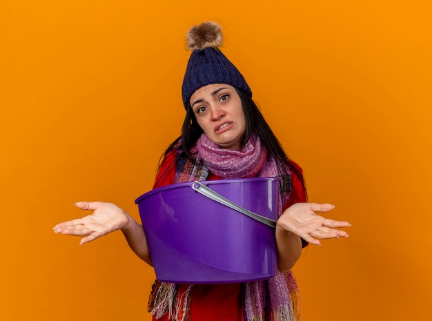 Desorientado joven enferma con gorro de invierno y bufanda sosteniendo un cubo de plástico con náuseas mirando al frente mostrando las manos vacías aisladas en la pared naranja