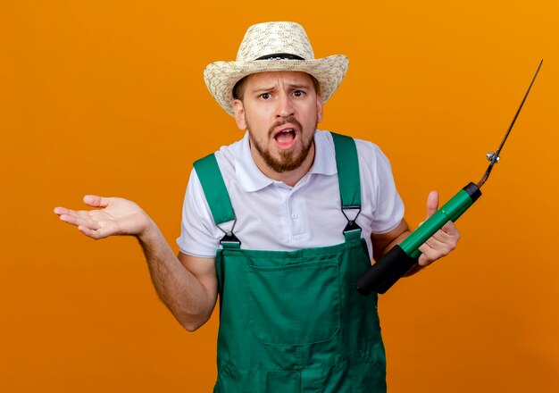 Desorientado joven jardinero eslavo guapo en uniforme y sombrero sosteniendo podadoras mostrando mano vacía mirando aislado