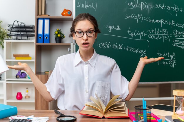 Desorientado joven profesora de matemáticas con gafas sentado en el escritorio con útiles escolares mirando al frente mostrando las manos vacías en el aula