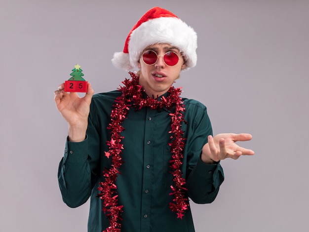 Desorientado joven rubio con gorro de Papá Noel y gafas con guirnalda de oropel alrededor del cuello sosteniendo el juguete del árbol de Navidad con fecha mirándolo mostrando la mano vacía aislada sobre fondo blanco