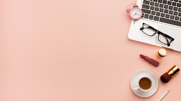 Despertador, anteojos en la laptop con taza de café y producto de maquillaje con fondo de durazno