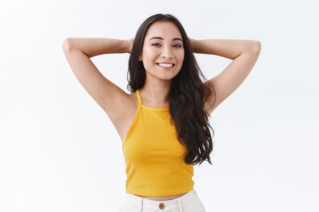 Despreocupada atractiva, despreocupada mujer del este de Asia que disfruta de vacaciones o fines de semana perezosos, tomados de la mano en pose relajada detrás de la cabeza, sonriendo alegremente, teniendo tiempo libre de pie en la pared blanca