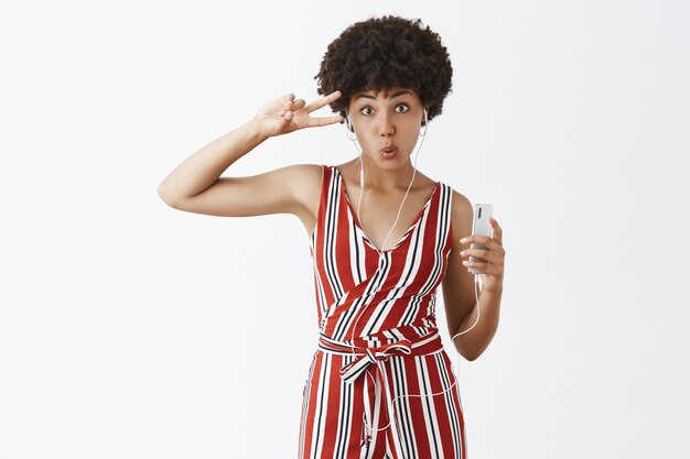 Despreocupada elegante mujer afroamericana de moda con cabello rizado escuchando música en auriculares sosteniendo smartphoen y mostrando discoteca o signo de la paz, doblando los labios, divirtiéndose