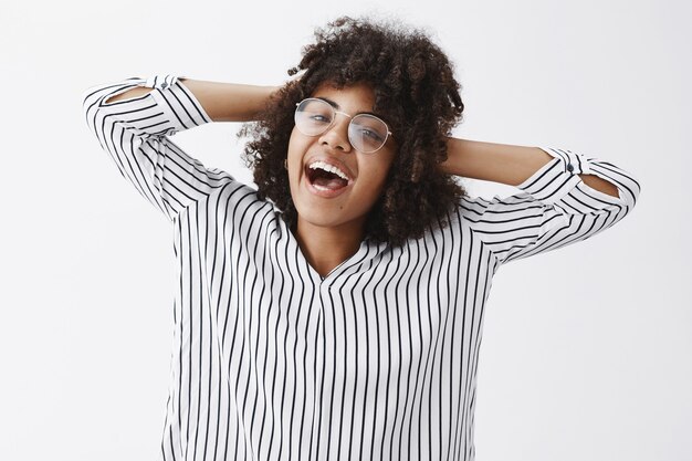 despreocupada, feliz y alegre, guapa mujer de piel oscura con peinado afro tomados de la mano detrás de la cabeza y sonriendo complacido y relajado llevando un estilo de vida elevado