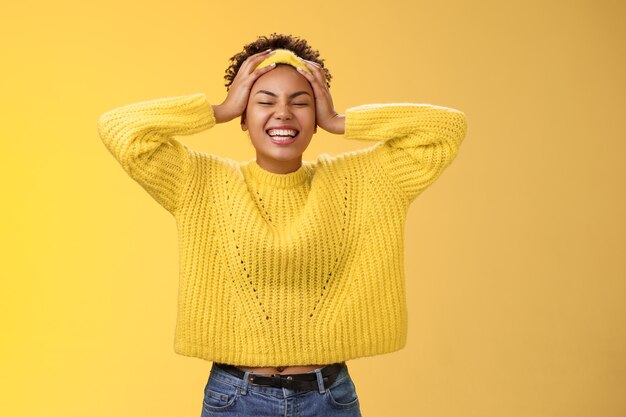 Despreocupada feliz niña africana afortunada en suéter con diadema tocando la cabeza divirtiéndose sonriendo aliviado emocionado riendo en voz alta cerrar los ojos regocijándose pasar un buen rato de fiesta, de pie fondo amarillo.