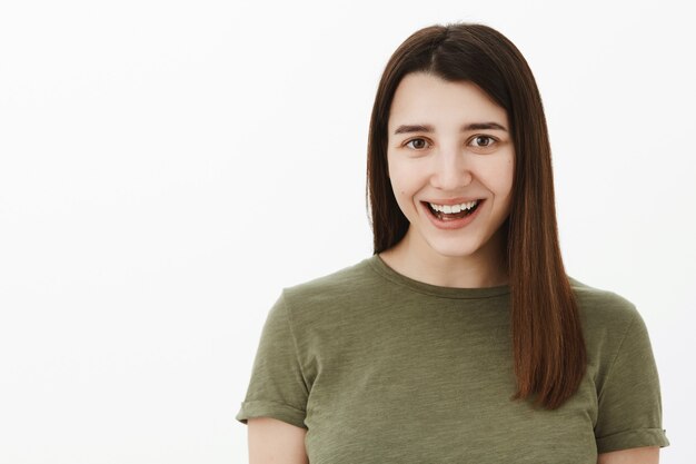 Despreocupada, feliz y optimista joven morena de unos 20 años con camiseta verde oliva sonriendo ampliamente con asombro y alegría, divirtiéndose, sintiéndose optimista y entretenida mientras está parado sobre una pared gris