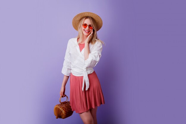 despreocupada mujer morena en vestido rosa de moda y sombrero de paja con bolsa de bambú en violeta.