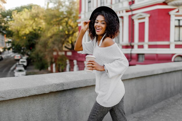 Despreocupada mujer negra con elegantes pelos afro y sombrero negro caminando en la ciudad y sosteniendo la taza de bebida. Atuendo casual. Perfecta sonrisa sincera. Divirtiéndose.