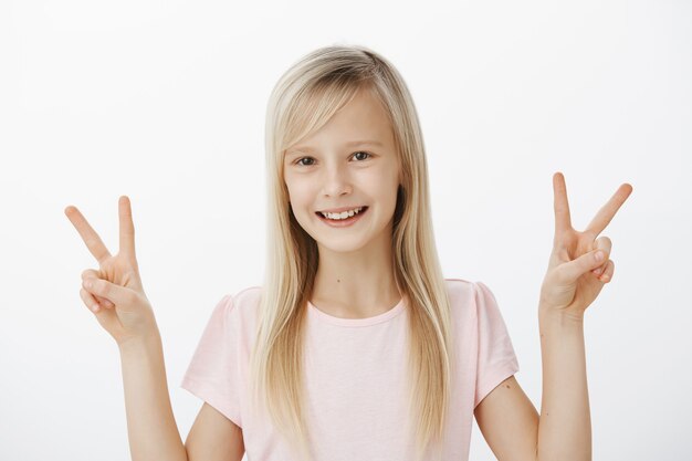 despreocupada niña positiva con cabello rubio en traje casual, mostrando victoria o gestos de paz con ambas manos y sonriendo alegremente, de pie contra la pared gris feliz