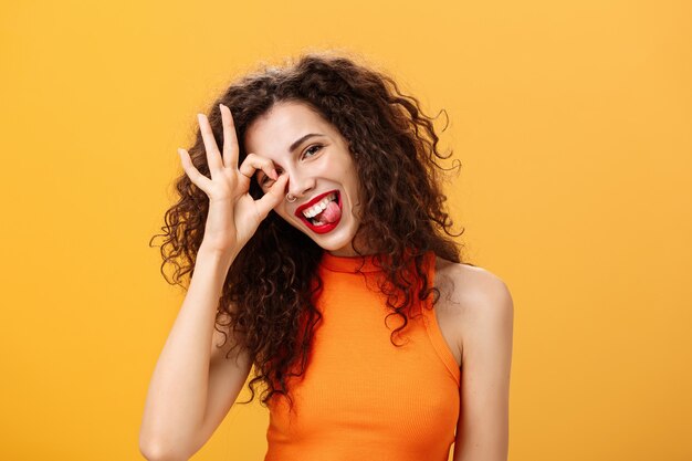 Despreocupada, optimista y enérgica mujer guapa con peinado rizado inclinando la cabeza y sacando la lengua juguetonamente mostrando bien o excelente signo sobre los ojos posando cerca de un fondo naranja.