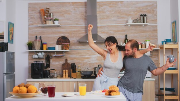 Despreocupada pareja feliz y alegre bailando y cantando en la cocina en la mañana de synny. Marido y mujer alegres riendo, cantando, bailando escuchando meditando, viviendo felices y sin preocupaciones. Gente positiva