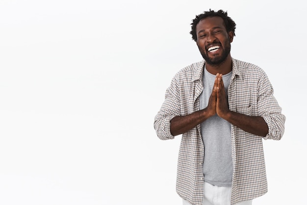 Foto gratuita despreocupado, feliz y agradecido, un hombre sonriente afroamericano con barba, peinado afro, junta las manos en oración y ríe, mostrando gratitud, agradeciendo la ayuda, de pie, fondo blanco.