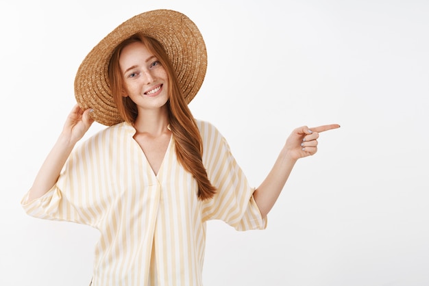 Despreocupado relajado atractivo pelirrojo femenino de vacaciones disfrutando de la cálida brisa de verano sosteniendo el sombrero de paja en la cabeza inclinando la cabeza y sonriendo alegremente apuntando a la derecha