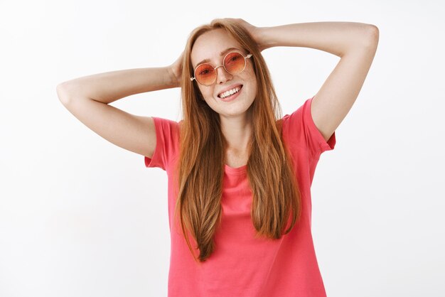 Despreocupado relajado y feliz joven pelirroja con gafas de sol de moda y camiseta rosa cogidos de la mano detrás de la cabeza